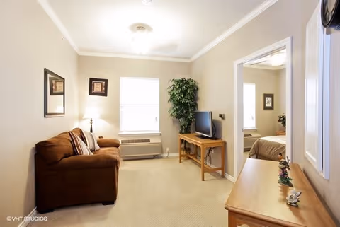 A cozy living room with a brown sofa, a small wooden TV stand holding a flat-screen television, a potted plant in the corner, a window with blinds, and framed artwork on the walls. Adjacent to the living room is a bedroom visible through an open doorway.