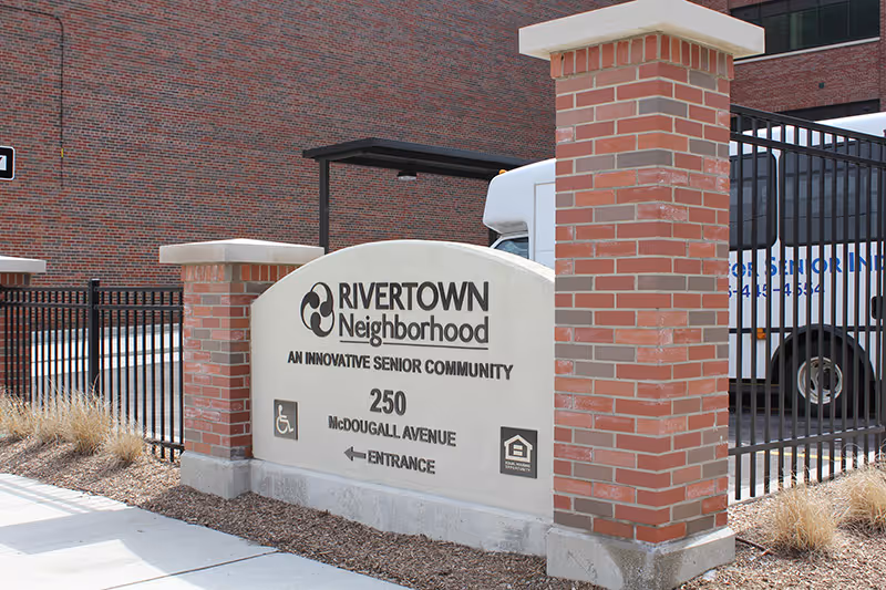 Outdoor stone sign reading 'RIVERTOWN Neighborhood' between brick pillars in front of a brick building with a parked van behind.