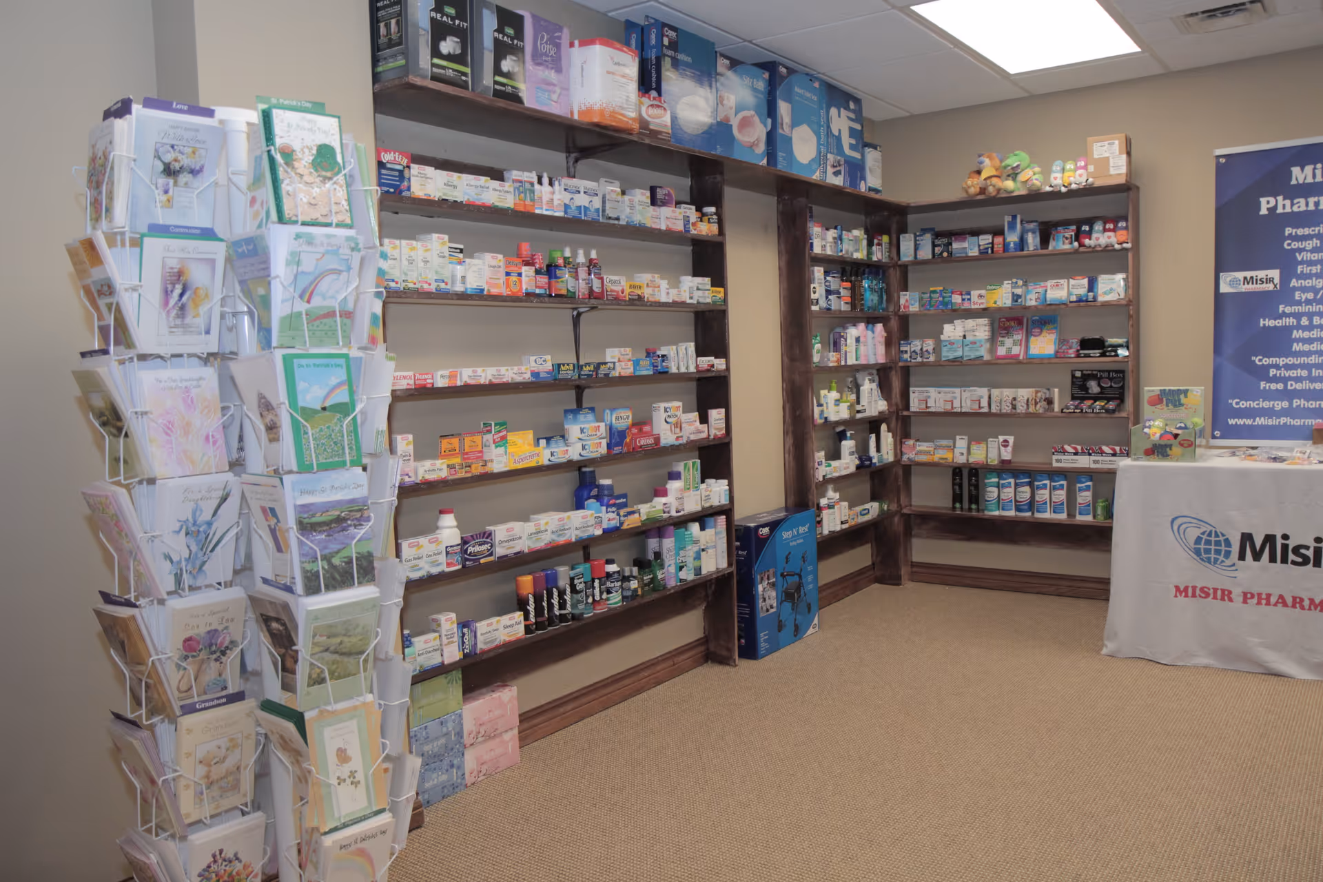 Interior view of a pharmacy area with shelves stocked with various medications, health products, and medical supplies. A rotating rack with greeting cards is positioned on the left side. A table covered with a cloth displaying the Misir Pharmacy logo is on the right side, along with a banner listing pharmacy services.
