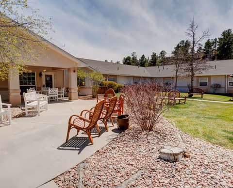 Outdoor patio area at Lark Springs facility with several wooden chairs arranged along a concrete pathway. The patio is adjacent to a single-story building with a covered porch area furnished with white chairs and tables. The surrounding landscape includes grass, small bushes, trees, and a rock bed.