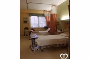Two hospital-style beds in a senior care room separated by a privacy curtain, with a window and bedside equipment.