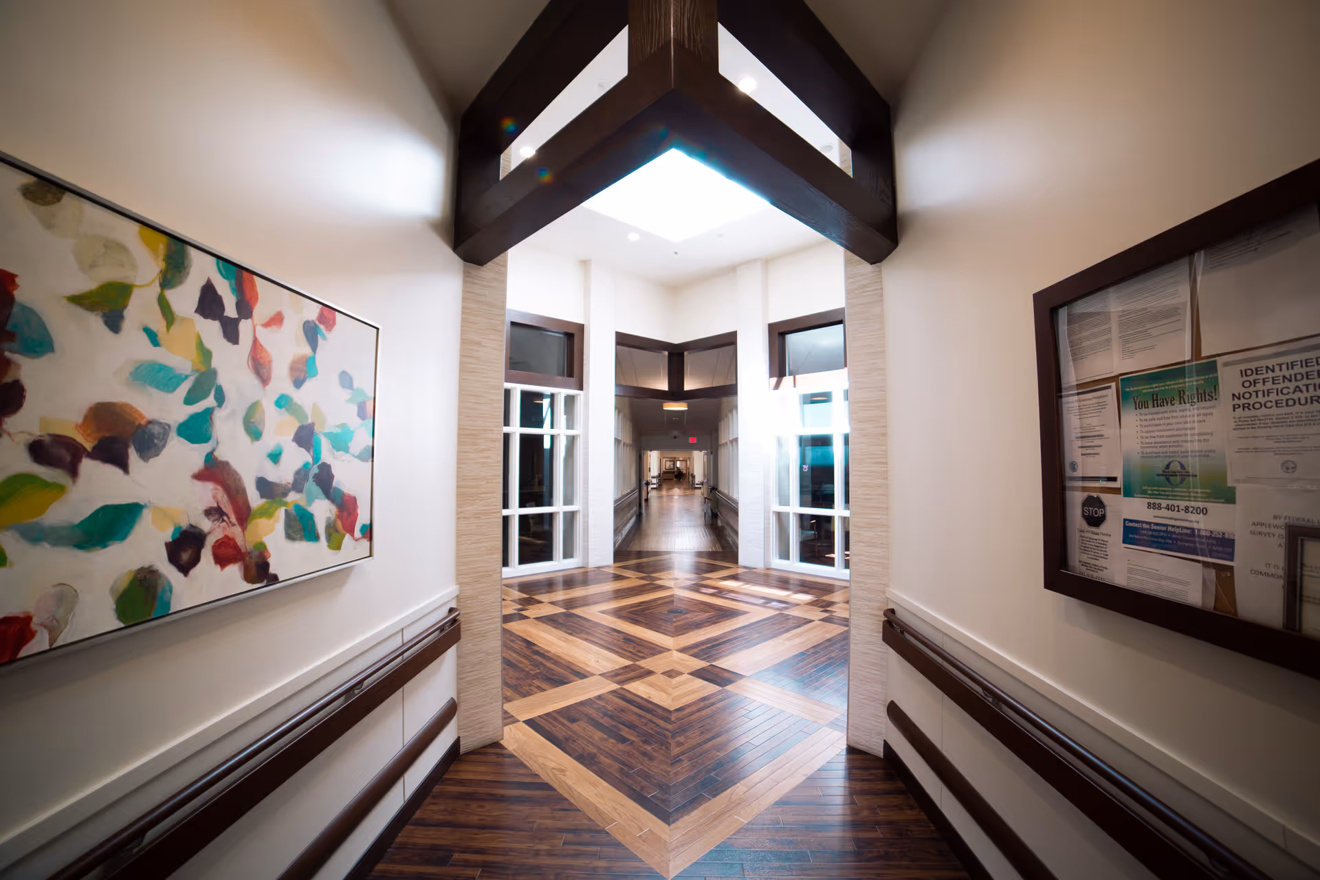 Bright, modern corridor in a senior living facility with patterned wood floors, handrails, colorful artwork on the left and a bulletin board on the right.
