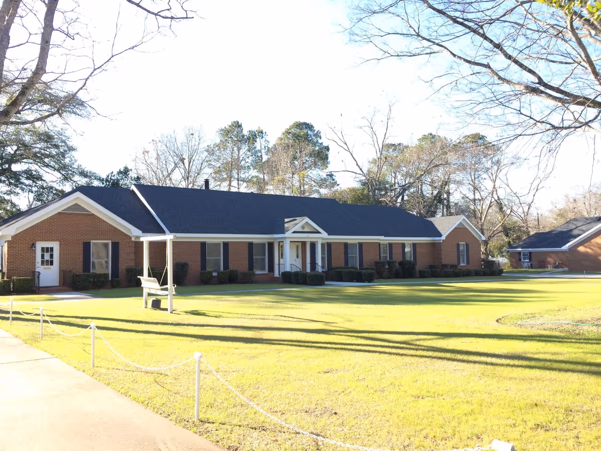 A single-story brick building with a dark roof, multiple windows with black shutters, and a white front door with a small porch. The building is surrounded by a well-maintained lawn with a swing bench on the left side and several leafless trees in the background under a clear sky.