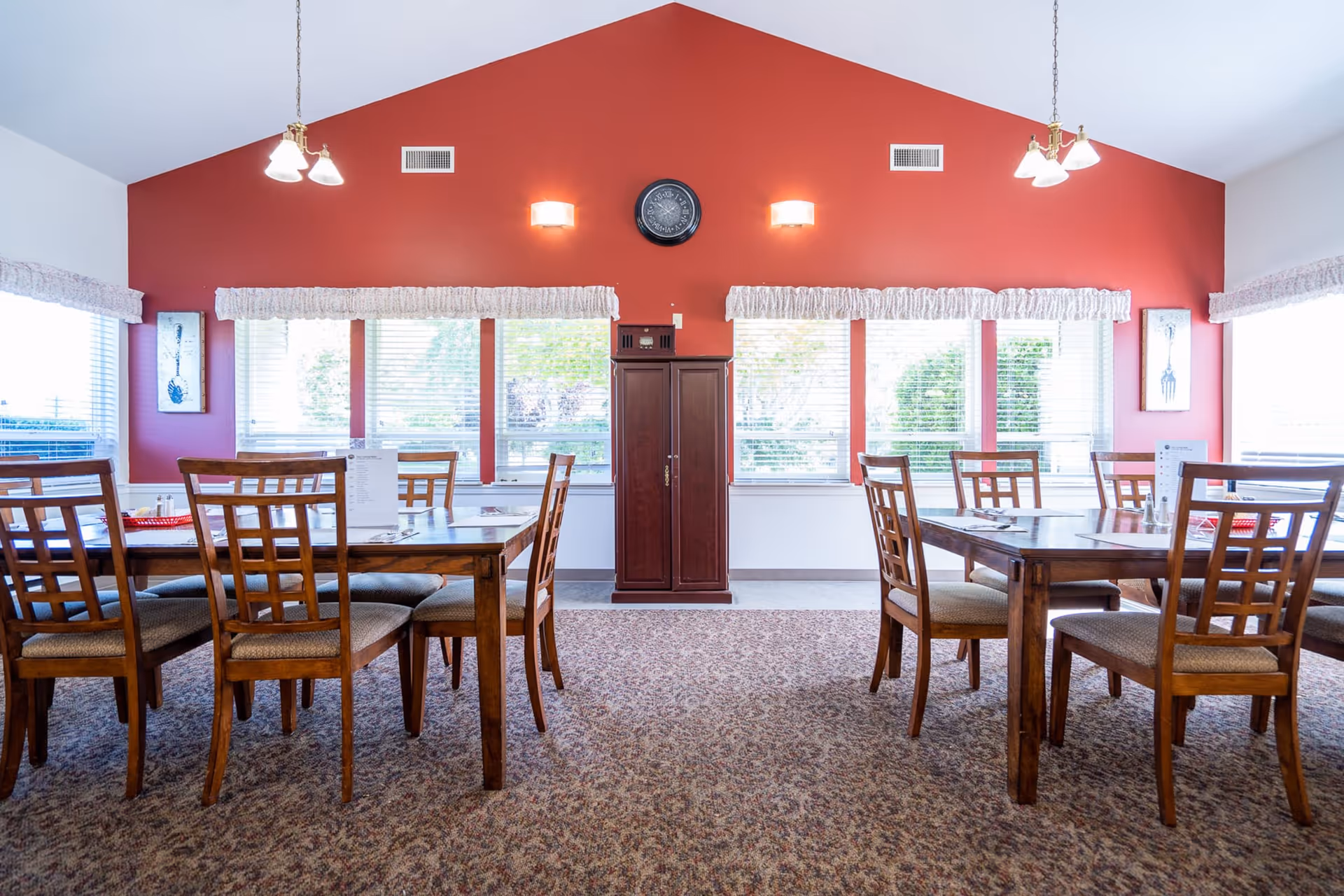 A bright dining room with wooden tables and chairs arranged neatly on a patterned carpet. The walls are painted red with white trim, and large windows with white blinds and valances let in natural light. Two hanging light fixtures illuminate the room, and a clock is mounted on the red wall above a wooden cabinet.