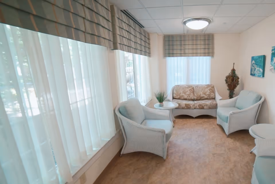 A bright sitting area with large windows covered by sheer white curtains and plaid valances. The room contains white wicker furniture including two armchairs with light blue cushions, a loveseat with floral cushions, and two small round tables. The walls are light-colored and decorated with two small paintings and a decorative wooden piece in the corner.