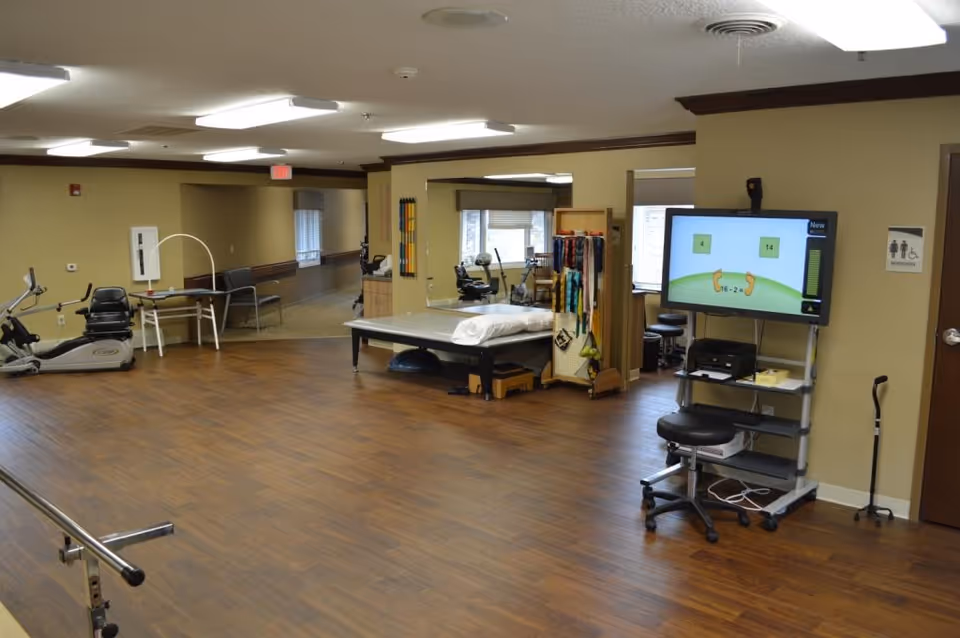 A spacious therapy or rehabilitation room with wooden flooring, exercise equipment including a recumbent bike, a treatment table with a pillow, a large mirror, and a TV screen displaying a math exercise. There is a stool, a cane leaning against the wall, and a sign indicating a restroom nearby.