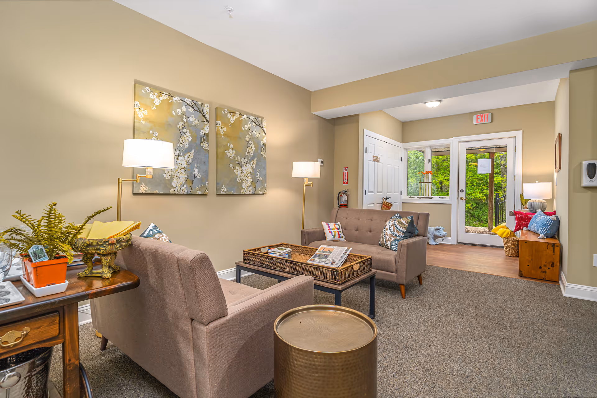 A cozy living room area with two beige sofas facing each other, a coffee table with a woven tray, and two floor lamps. The walls are painted light beige and decorated with two floral paintings. There is a small wooden side table with a plant and other items on the left. In the background, there is a door with glass panels leading outside, a wooden bench with colorful cushions, and a fire extinguisher mounted on the wall.