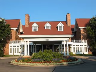 Front exterior view of a large brick building with a red roof, white columns at the entrance, and a circular driveway with a landscaped flower bed in front.