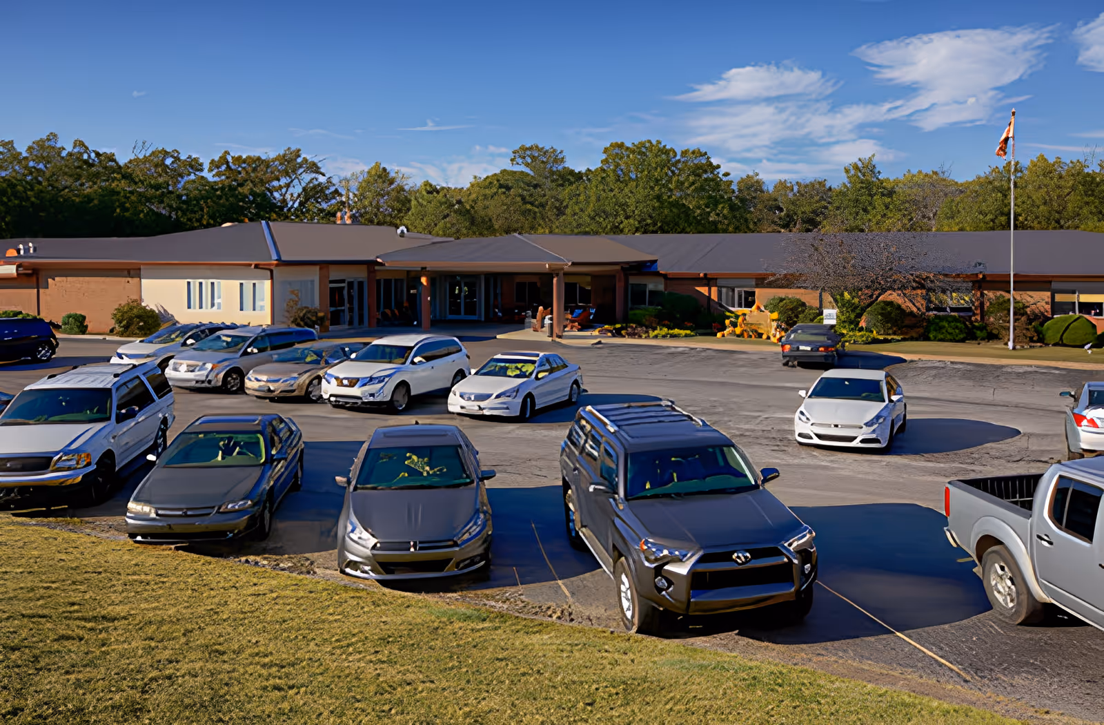 Exterior view of AHC Savannah senior living facility with a parking lot in front. Several cars are parked, and the building is a single-story structure with a covered entrance. Trees and a flagpole are visible in the background under a partly cloudy sky.
