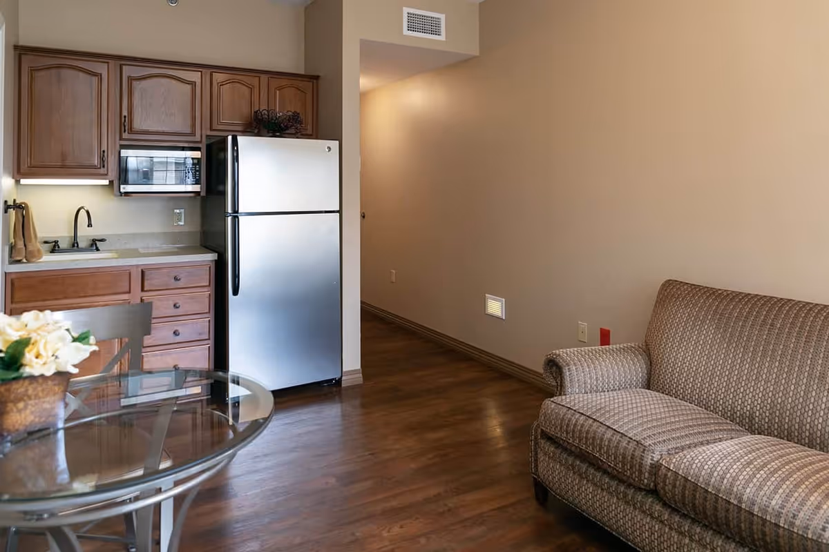 Interior view of a senior living facility apartment showing a small kitchen area with wooden cabinets, a stainless steel refrigerator, a microwave, and a sink. Adjacent to the kitchen is a glass-top dining table with chairs and a patterned upholstered sofa on the right side. The floor is wooden, and the walls are painted beige.