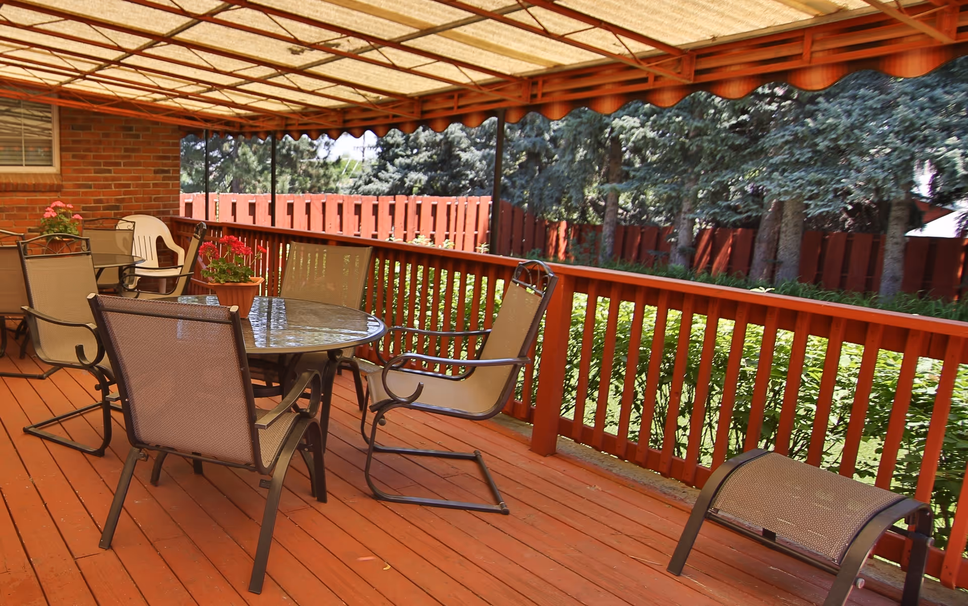 Covered outdoor patio area with a glass-top round table surrounded by four metal mesh chairs. There are potted plants with red flowers on the table and another table with chairs in the background. The patio has a wooden floor and railing painted in reddish-brown, with a red wooden fence and green trees visible beyond the railing.