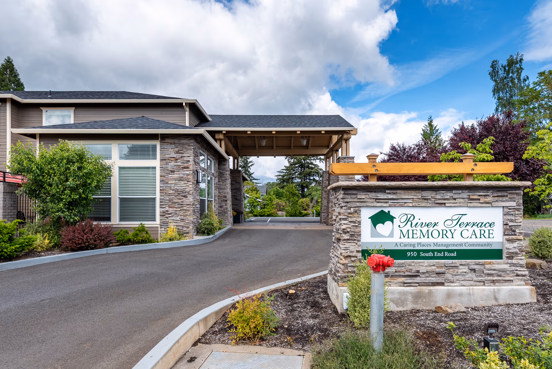 Entrance driveway to River Terrace Memory Care facility with a stone sign displaying the facility name and address. The building has stone and beige siding with large windows, surrounded by landscaped greenery and trees under a partly cloudy sky.