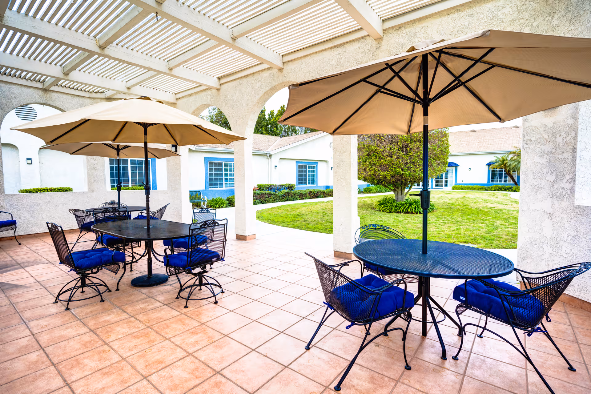 Outdoor patio area with round metal tables and chairs with blue cushions, shaded by large beige umbrellas. The patio has a tiled floor and is partially covered by a pergola. In the background, there is a grassy courtyard with bushes and a tree, surrounded by white buildings with blue-trimmed windows.