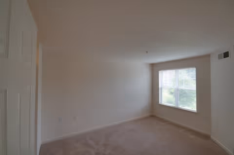 Empty carpeted bedroom with beige walls and a window on the right letting in natural light.