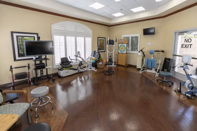 A rehabilitation room with exercise equipment including stationary bikes, a recumbent bike, and strength training machines. The room has wooden flooring, large windows with white shutters, a TV mounted on a stand, and a 'NO EXIT' sign on a glass door.