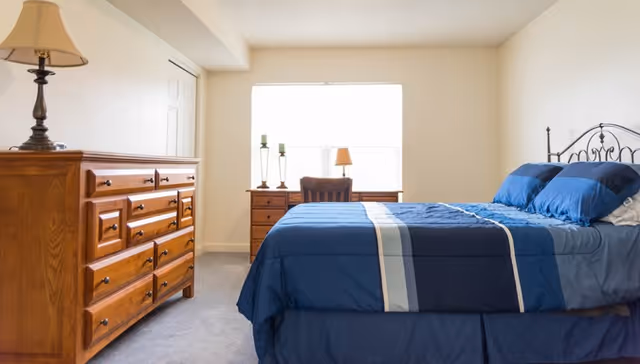 Bright bedroom with a blue-bedspread double bed, wooden dresser, nightstand and a window letting in light.