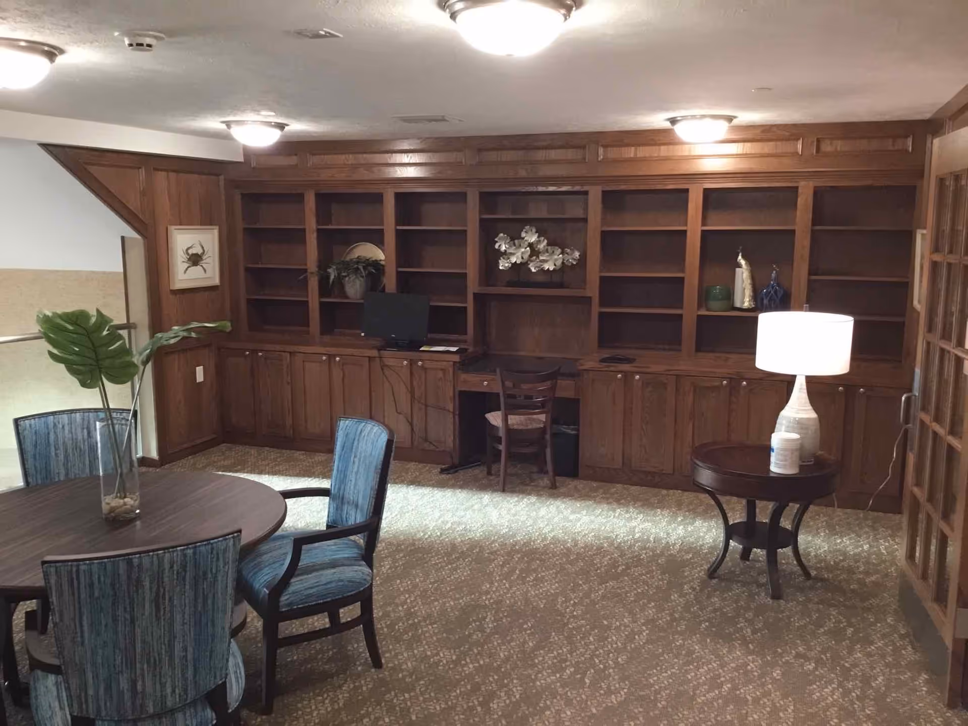 Cozy communal lounge with a round table and upholstered chairs, built-in wooden bookshelves and a small desk with a computer.