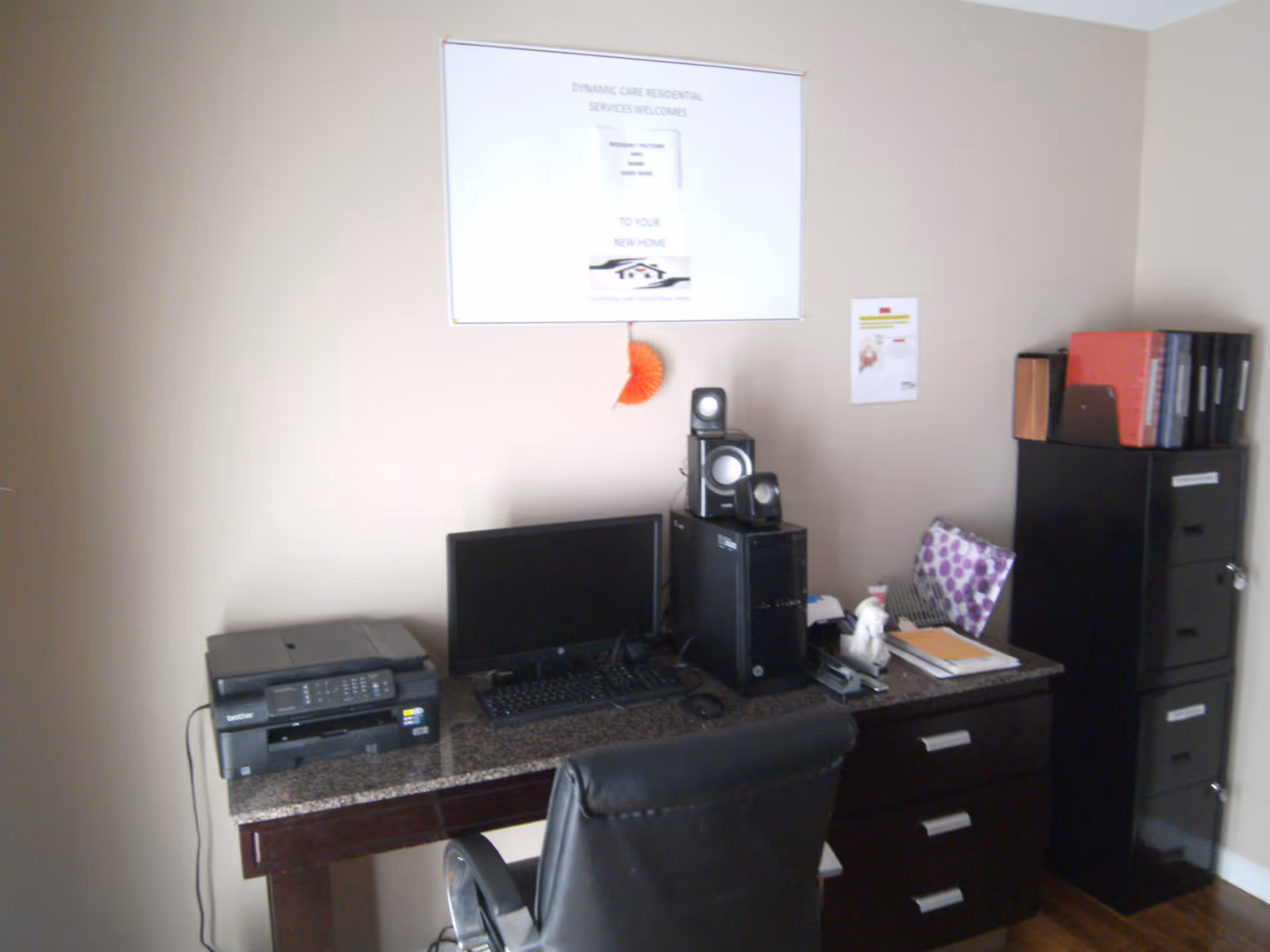 An office workspace with a computer monitor, keyboard, desktop tower with speakers on top, a printer, and various office supplies on a dark wooden desk. There is a black office chair in front of the desk and a black filing cabinet with binders on top to the right. A sign on the wall above the desk reads 'Dynamic Care Residential Services Welcomes You to Your New Home.'