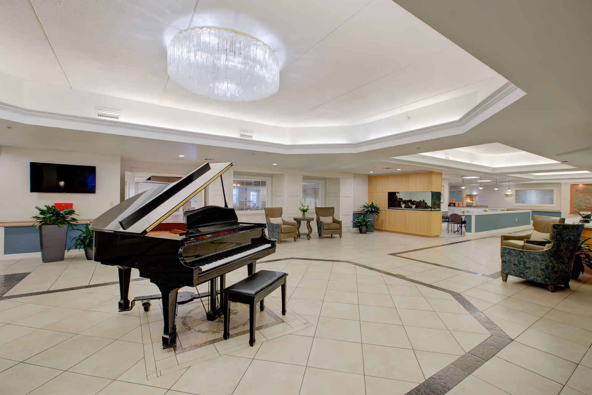 Spacious and well-lit common area featuring a black grand piano with a matching bench in the center. The room has tiled flooring with decorative patterns, comfortable armchairs, a large chandelier, and a built-in aquarium. There are plants and a mounted TV on the walls, creating a welcoming and elegant atmosphere.