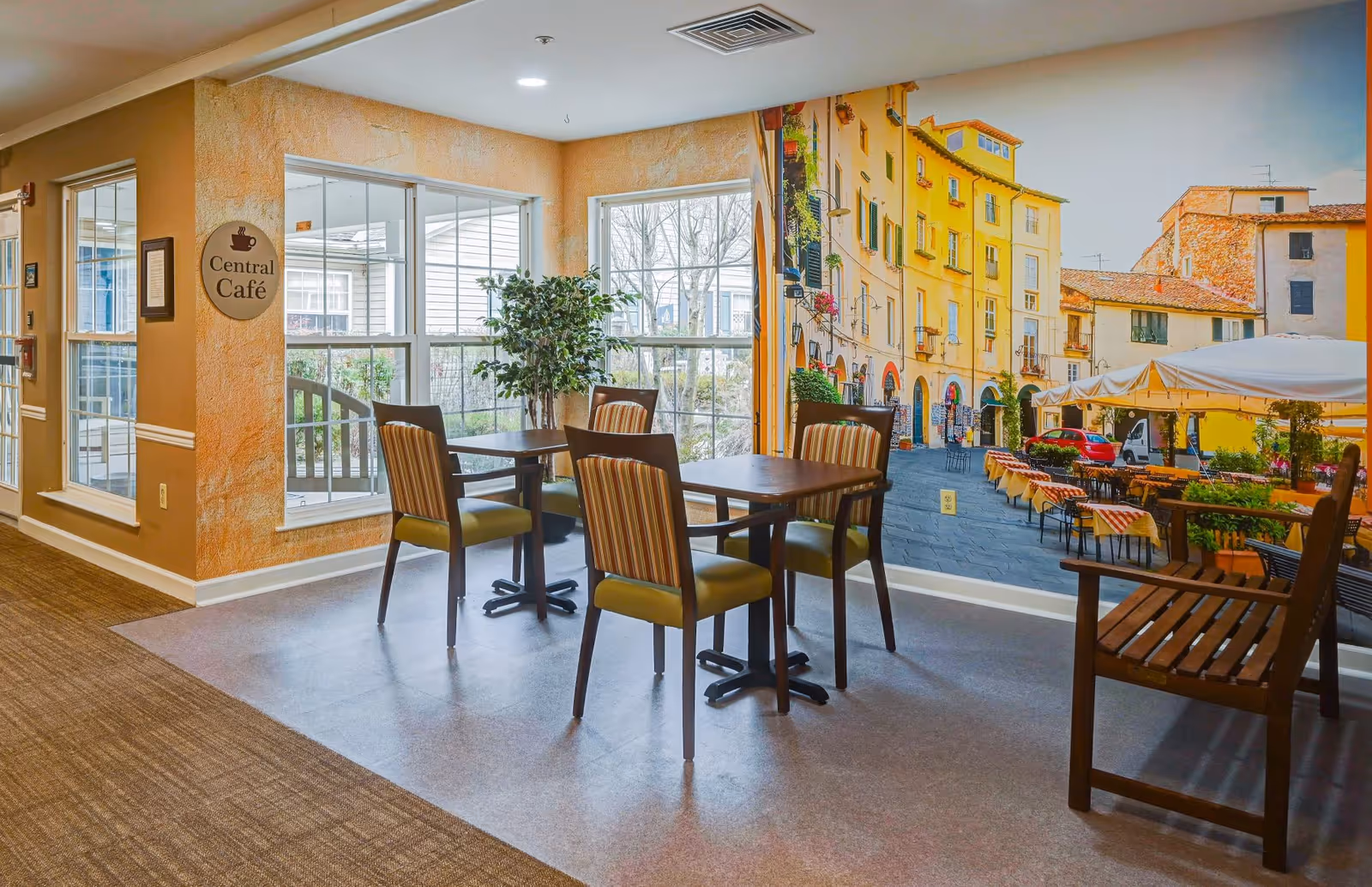A small dining area with two tables and four chairs, located next to large windows letting in natural light. The walls are painted warm beige, and one wall features a large mural of a European street scene with colorful buildings and outdoor café tables. A sign on the wall reads 'Central Café'.
