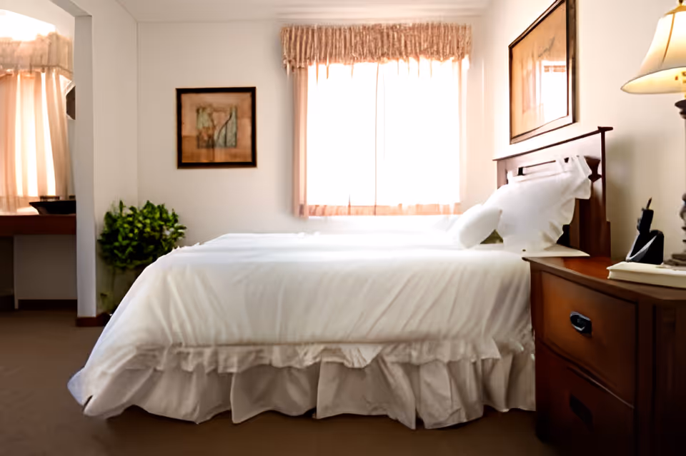 A neatly made bed with white bedding and pillows in a bedroom. There is a wooden nightstand with a lamp and some items on it to the right of the bed. A window with sheer curtains lets in natural light behind the bed. A framed picture hangs on the wall above the nightstand and another framed artwork is on the adjacent wall. A green potted plant is visible in the corner near an open doorway leading to another room with a vanity and mirror.
