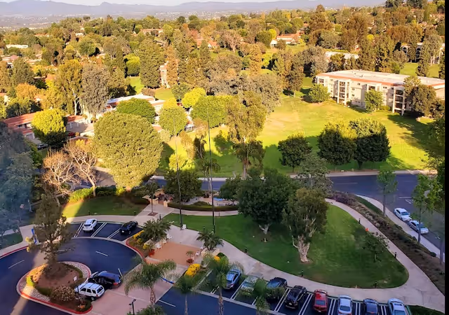 Aerial view of a senior living community with green lawns, trees, pathways, parked cars, and residential buildings in the background under a clear sky.
