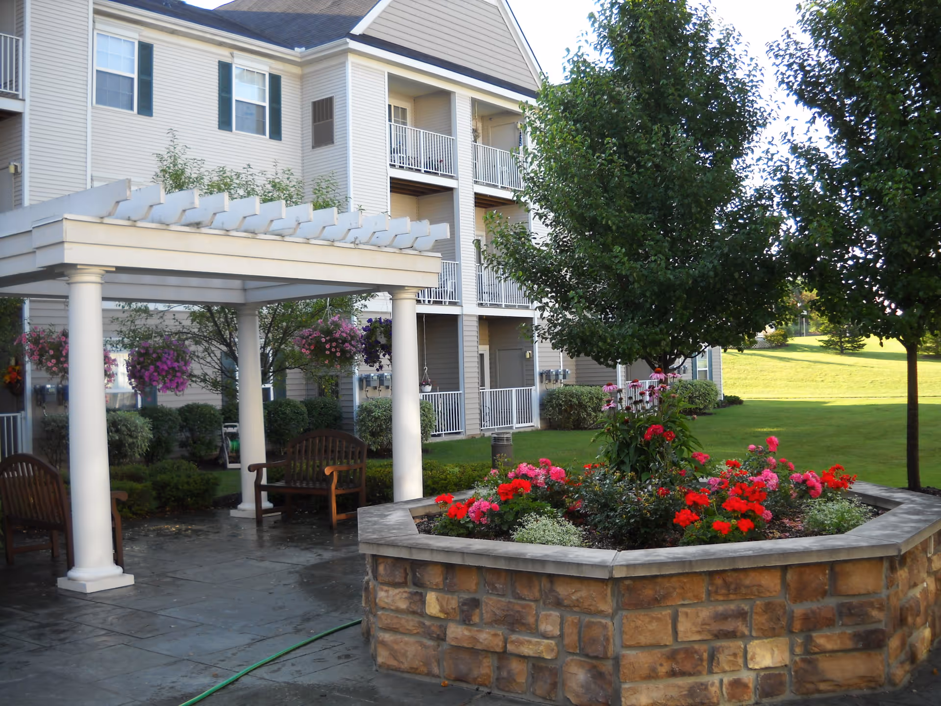 Outdoor garden area at a senior living facility with a stone planter filled with colorful flowers and a small tree. There is a white pergola with two wooden benches underneath, and a multi-story building with balconies in the background. The lawn is green and well-maintained.