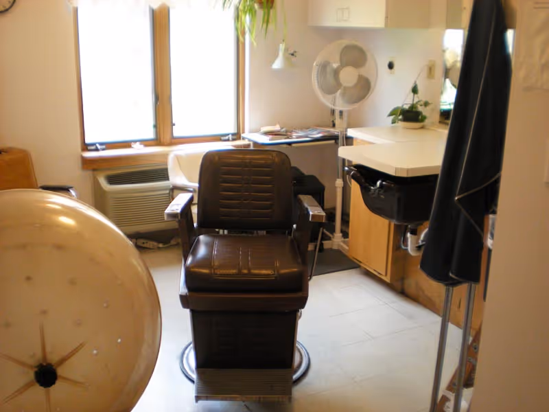 Interior view of a small salon or barber area with a vintage brown leather barber chair in the center, a hair dryer hood on the left, a white sink with a black basin on the right, a standing fan, a small table with magazines, and a window with natural light coming through.