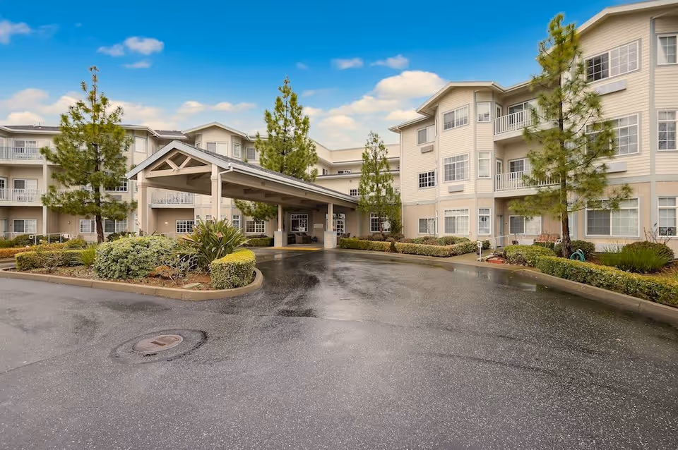 Exterior view of Country Crest Senior Living facility showing a three-story building with balconies, a covered entrance, landscaped bushes and trees, and a wet driveway under a partly cloudy sky.