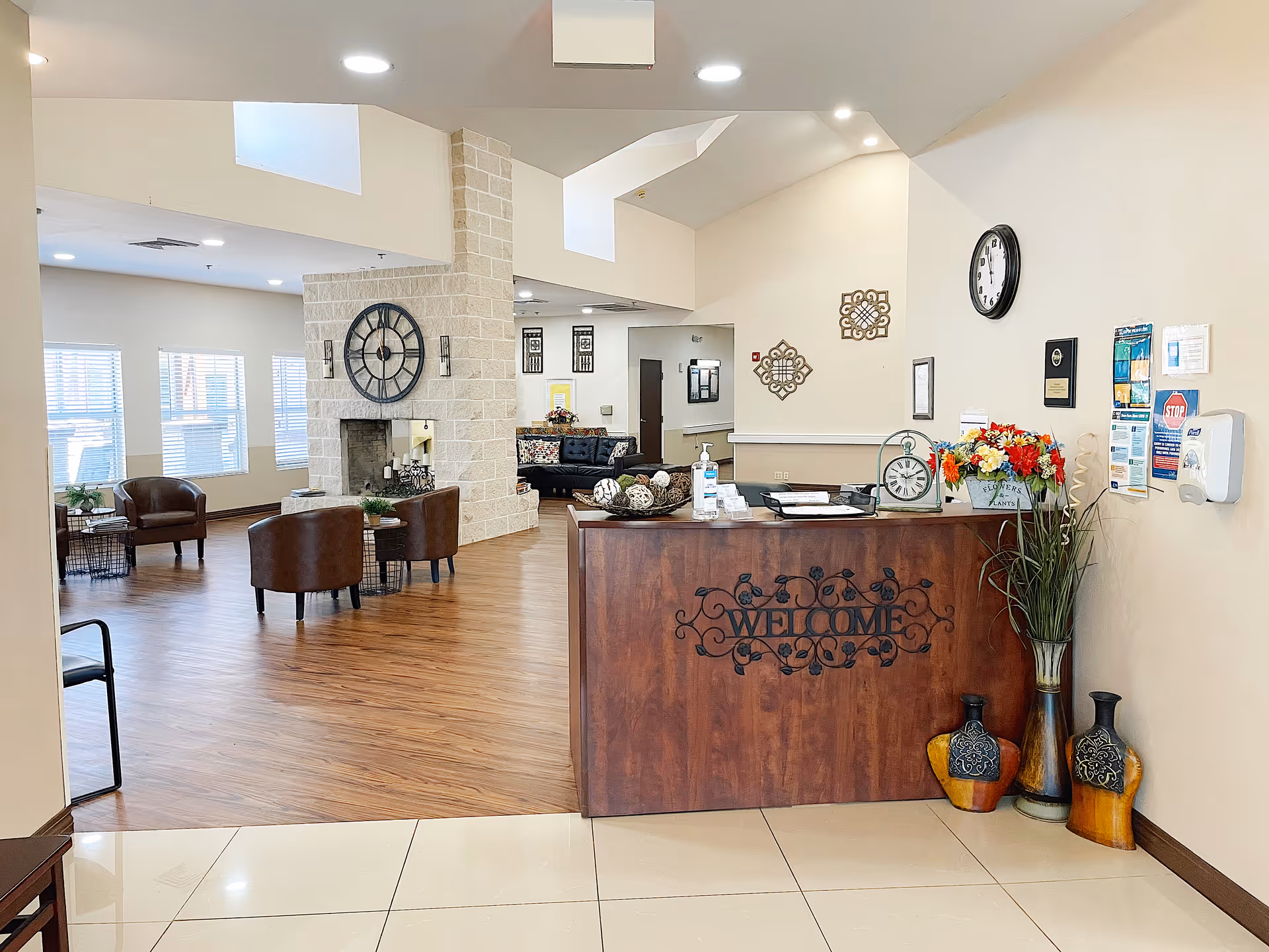 Interior view of a senior living facility reception area with a wooden welcome desk adorned with flowers, a clock, and decorative items. Behind the desk is a spacious lounge area with leather chairs, a large wall clock above a stone fireplace, and sofas. The room has wooden flooring and large windows letting in natural light.