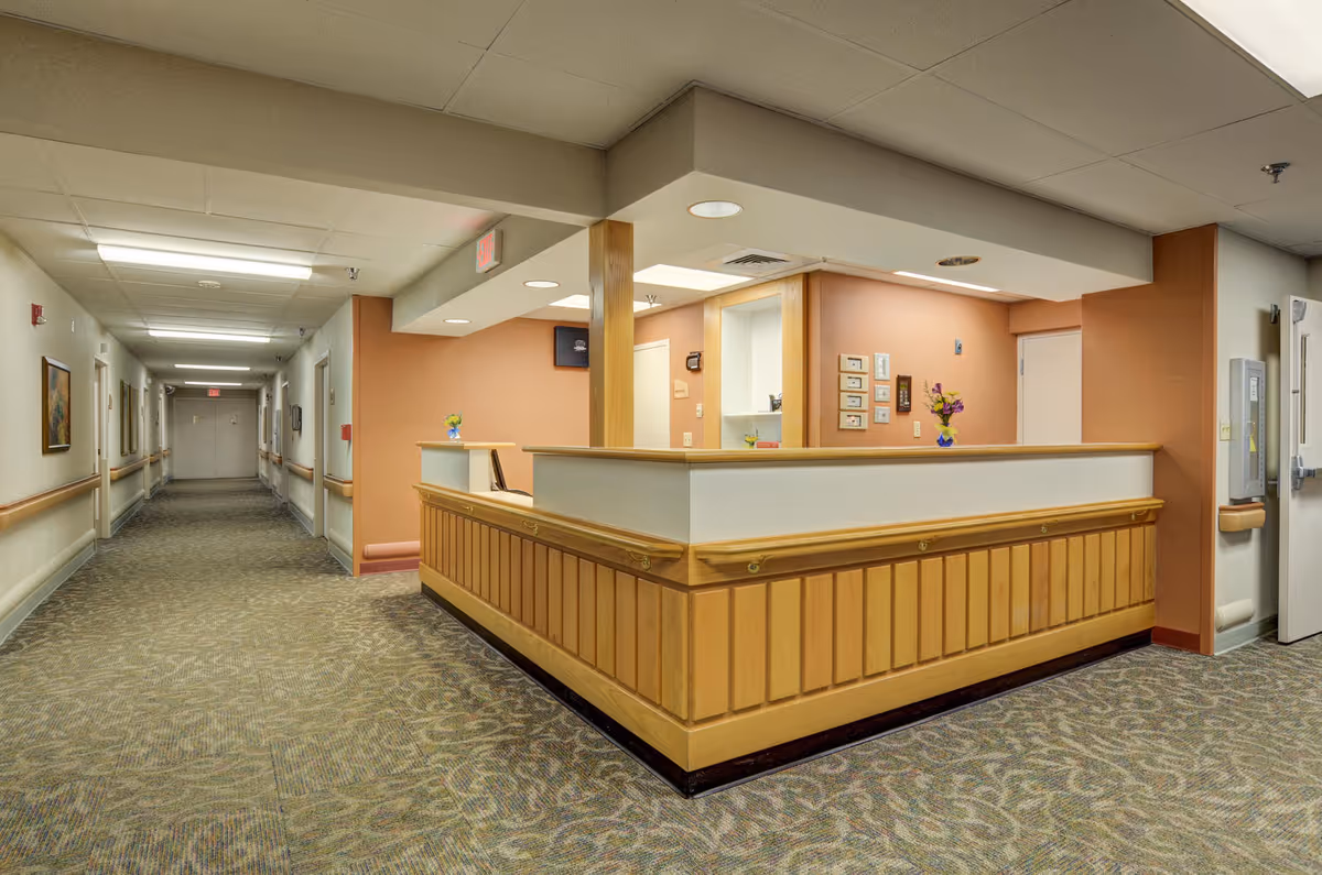 Interior view of a nursing facility hallway with a reception desk on the right side. The hallway is carpeted with patterned flooring and has handrails along the walls. The reception desk is wooden with a light-colored countertop and has small flower vases placed on it. The walls are painted in a warm beige and orange tone, and there are ceiling lights illuminating the area.