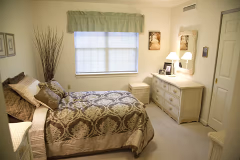 A cozy bedroom with a bed covered in a brown and beige patterned comforter and multiple pillows. There is a window with closed blinds and a green valance above it. To the right, there is a wicker dresser with a mirror above it, a lamp, and framed photos. A wicker nightstand is next to the dresser, and a door is visible on the right side of the room. The walls are painted light beige.