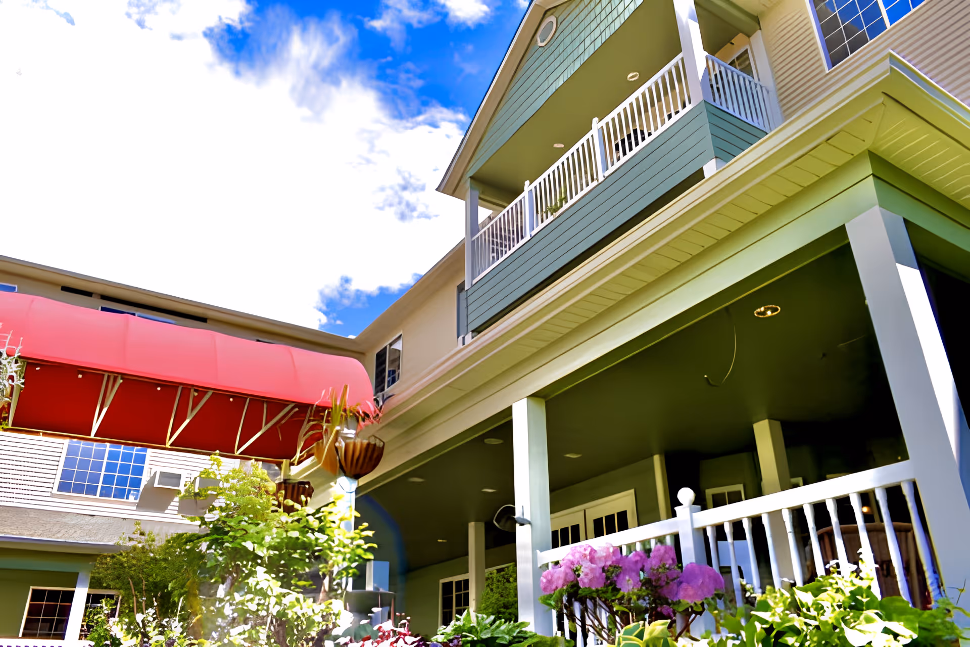 Exterior view of a senior living facility building with green siding, white railings on balconies, a red awning, and lush greenery including flowering plants in the foreground under a partly cloudy blue sky.