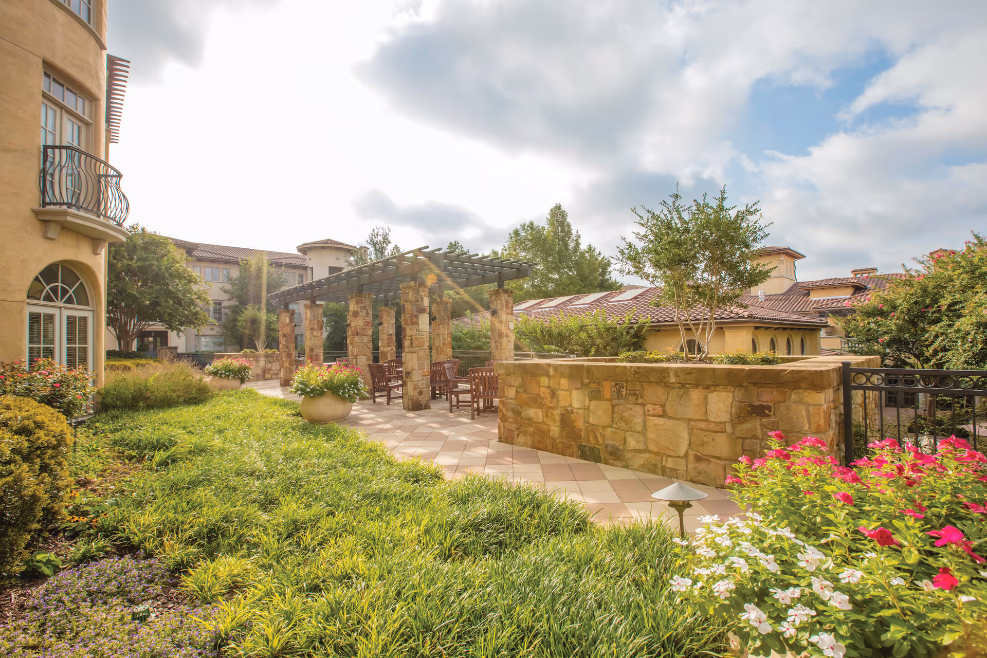 Outdoor garden area at a senior living facility with green grass, colorful flowers, stone pillars supporting a pergola, wooden benches, and a stone wall. The building with arched windows and balconies is visible in the background under a partly cloudy sky.