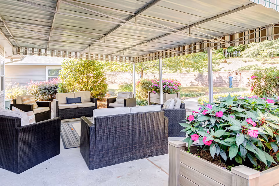 Covered outdoor patio area with black wicker furniture including sofas and chairs with cushions, surrounded by planters with pink flowers and greenery, and a striped awning overhead.