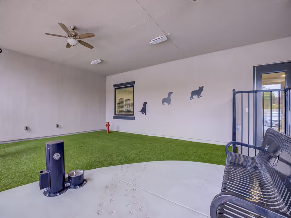 Covered outdoor pet area with artificial turf, a pet water station, bench, and dog silhouette wall decals on the wall.