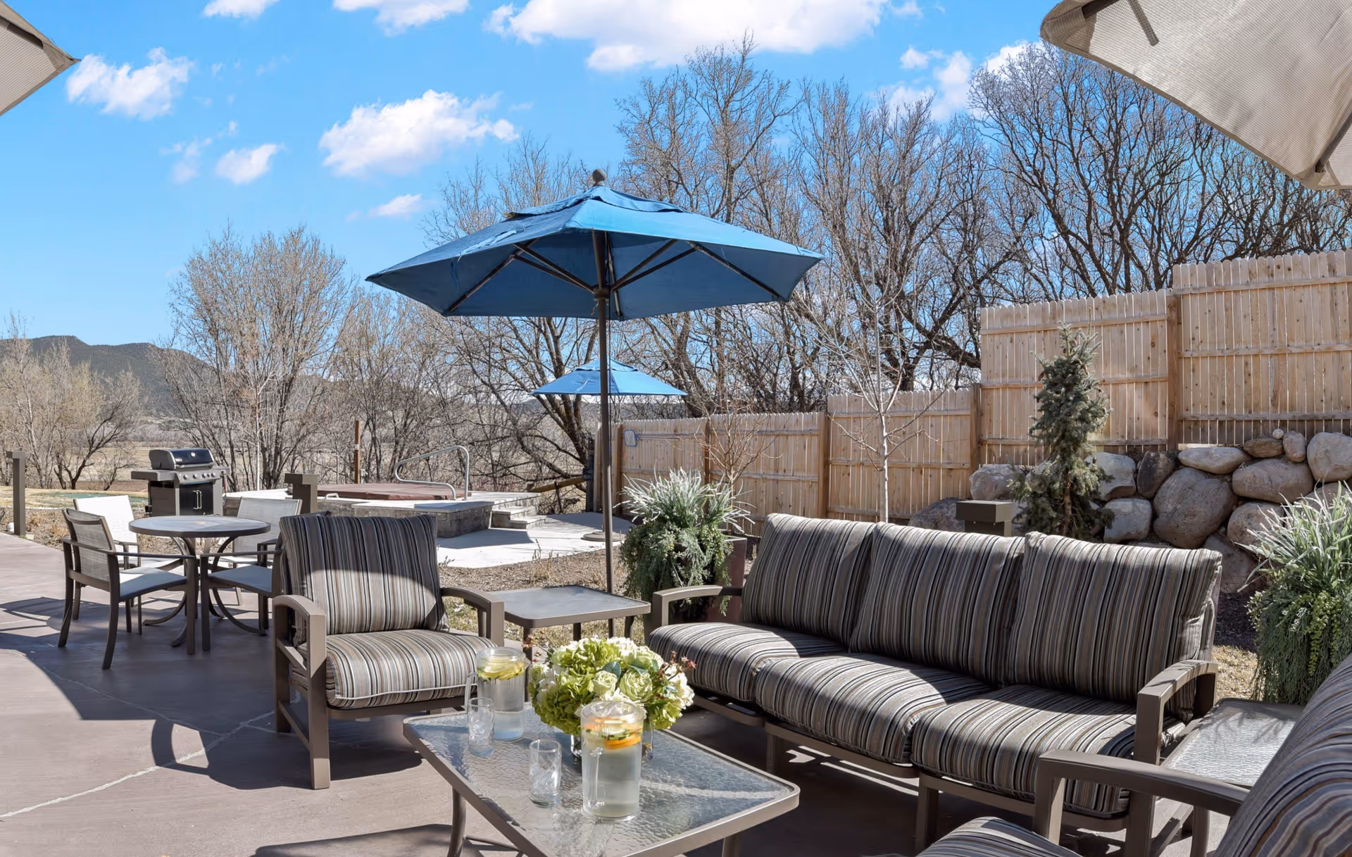 Outdoor patio area at Sopris Lodge at Carbondale Senior Living featuring cushioned seating with striped upholstery, glass-top tables with pitchers of water and glasses, blue umbrellas providing shade, a barbecue grill, and a hot tub. The area is surrounded by a wooden fence and leafless trees under a clear blue sky.