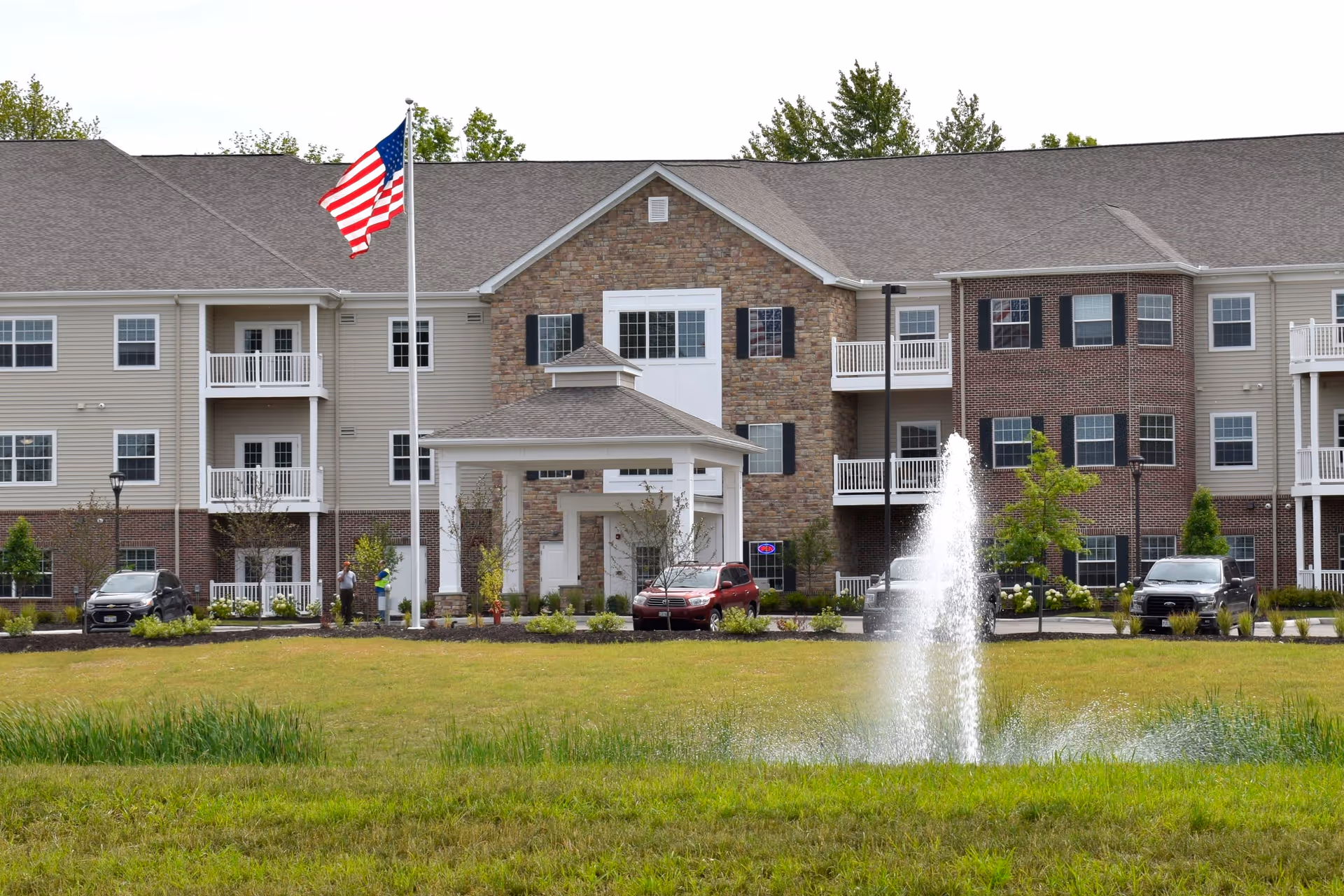 Front exterior view of a senior living facility named Springwood Hudson with a large American flag on a flagpole, a water fountain in the foreground, and several cars parked near the entrance.