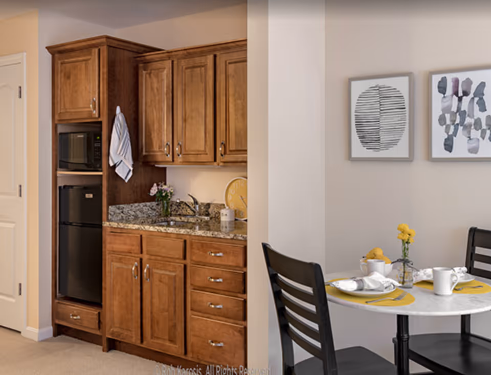 A small kitchen area with wooden cabinets, a granite countertop, a sink, a microwave, and a mini refrigerator. Adjacent to the kitchen is a round dining table set for two with black chairs, yellow placemats, white dishes, and a small vase with yellow flowers. Two abstract art pieces hang on the wall above the dining table.