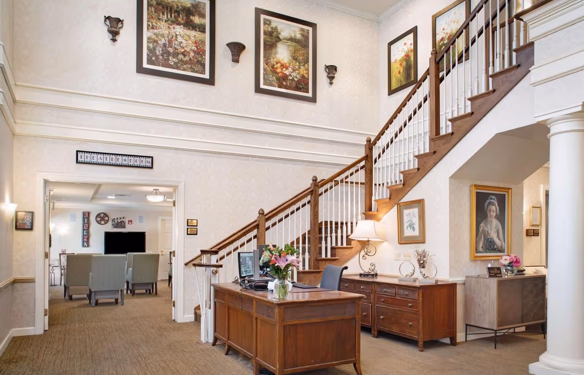 Interior view of a senior living facility lobby area with a wooden reception desk adorned with a vase of flowers, a staircase with wooden handrails and white balusters, framed paintings on the walls, and a doorway leading to a theater room with chairs and a large TV screen.