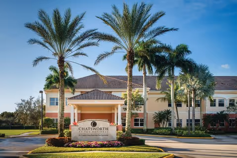 Front exterior view of Chatsworth Assisted Living Community at PGA National building with palm trees and a clear blue sky.