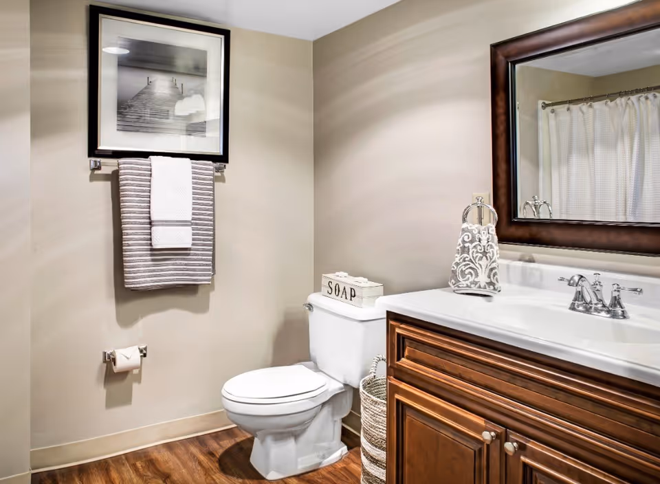 A clean bathroom with a white toilet, wooden vanity with a white countertop and silver faucet, a large framed mirror above the sink, a towel rack with striped and white towels, a basket beside the toilet, and a framed black and white photo of a pier on the wall.