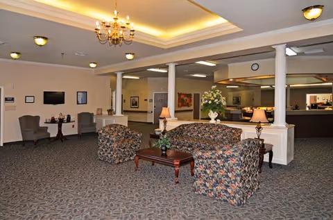A spacious senior living facility common area with floral patterned armchairs and a matching sofa arranged around a wooden coffee table. The room features a carpeted floor, white columns, and a ceiling with recessed lighting and a chandelier. There are two table lamps on side tables, a flower arrangement on the coffee table, and a reception desk in the background.