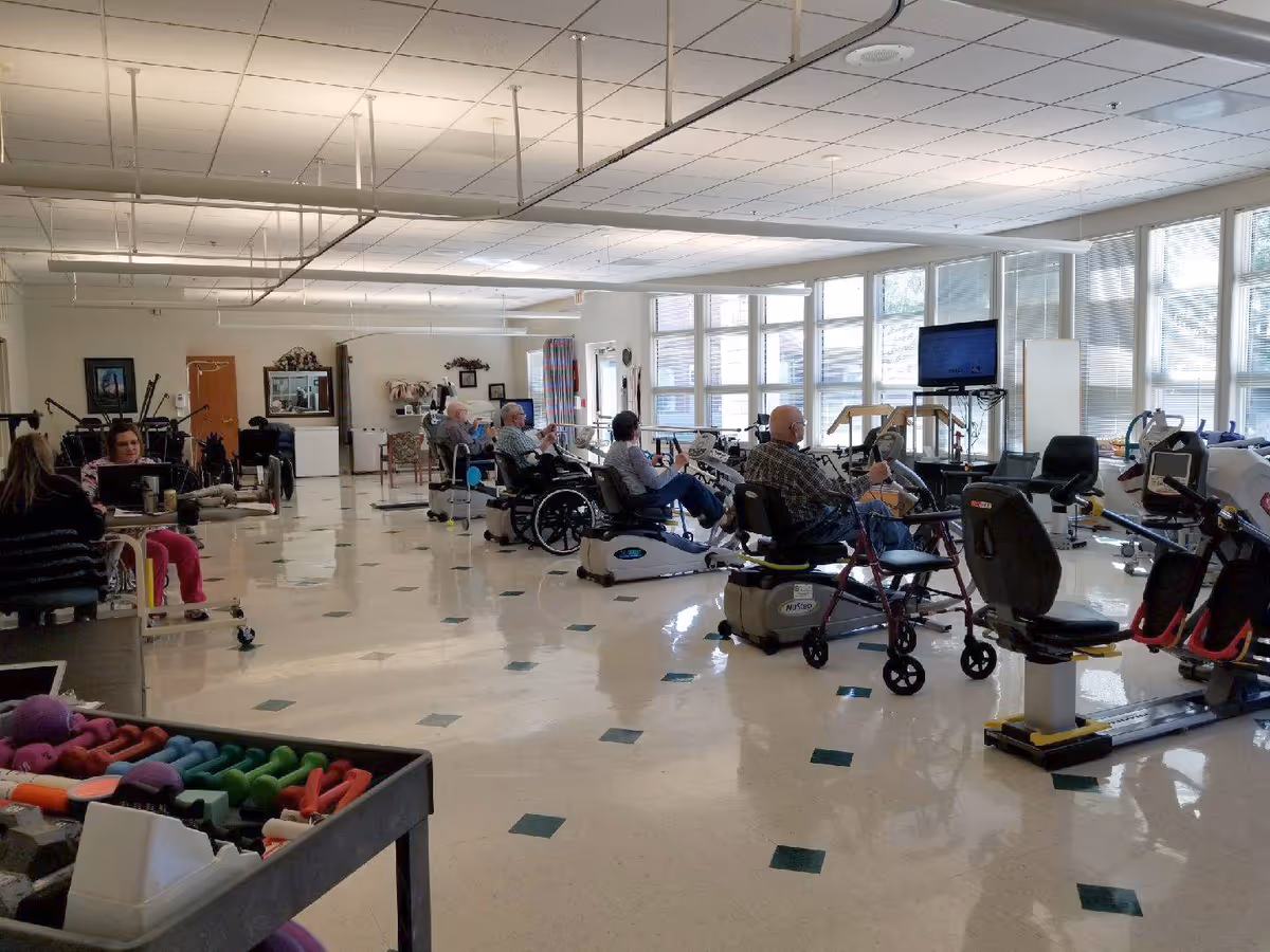 A spacious rehabilitation or therapy room with several elderly individuals using exercise equipment such as stationary bikes and other fitness machines. The room has large windows allowing natural light, a television mounted on the wall, and various exercise weights in the foreground. The floor is tiled with a pattern of green squares.