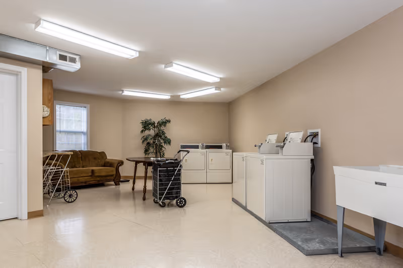 A laundry room in a senior living facility featuring several white washing machines and dryers along the walls, a utility sink on the right, a brown couch and a small table with a potted plant in the background, and laundry carts in the center of the room. The room has beige walls, a tiled floor, and fluorescent ceiling lights.