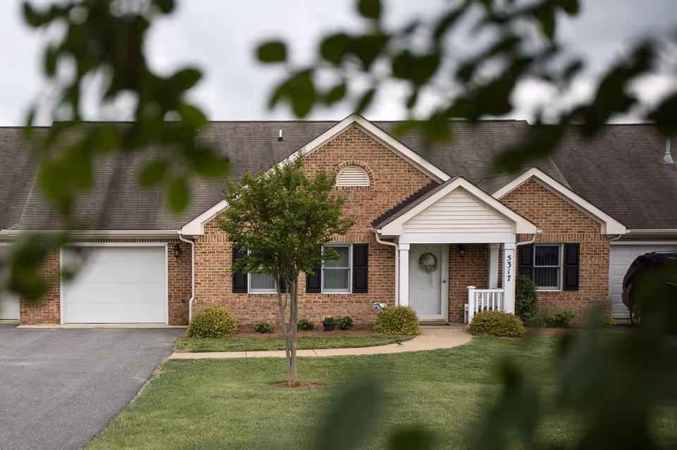 Brick single-story building with a central white porch entrance, attached garage, lawn and a small tree framed by blurred foreground leaves.