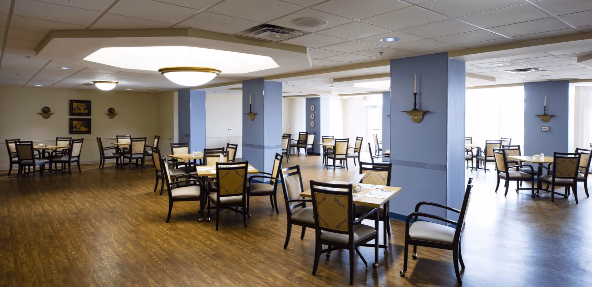 A spacious dining room in a senior living facility with multiple tables and chairs arranged neatly. The room has wooden flooring, blue pillars with candle holders, and soft lighting from ceiling fixtures. There are framed pictures on the far wall and large windows letting in natural light.