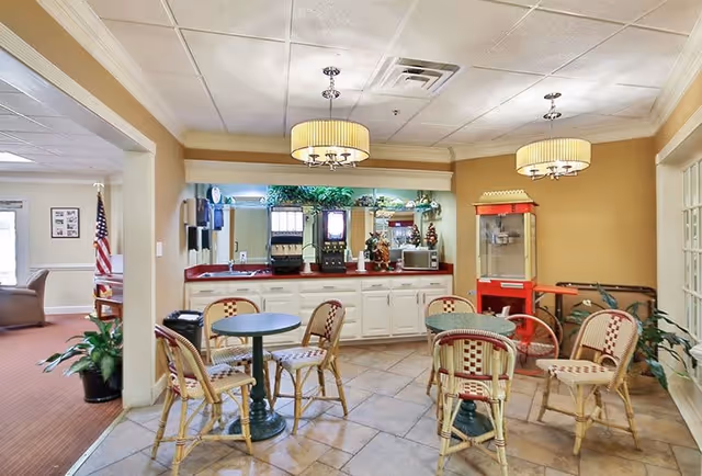 A small dining area with round tables and wicker chairs featuring red and beige patterns. In the background, there is a counter with beverage dispensers, a microwave, and a popcorn machine. The room has beige walls, tiled flooring, and two ceiling light fixtures with drum shades. A doorway leads to another room with carpeted flooring and an American flag visible.