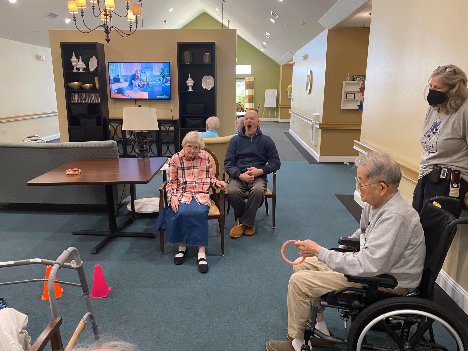 A group of elderly individuals in a common area of a senior living facility. One woman in a plaid shirt and blue skirt sits in a chair, a man next to her yawns, and another man in a wheelchair holds a ring. A staff member wearing a mask stands nearby. The room has a TV mounted on the wall, shelves with decorative items, and a table with some colorful rings on it.