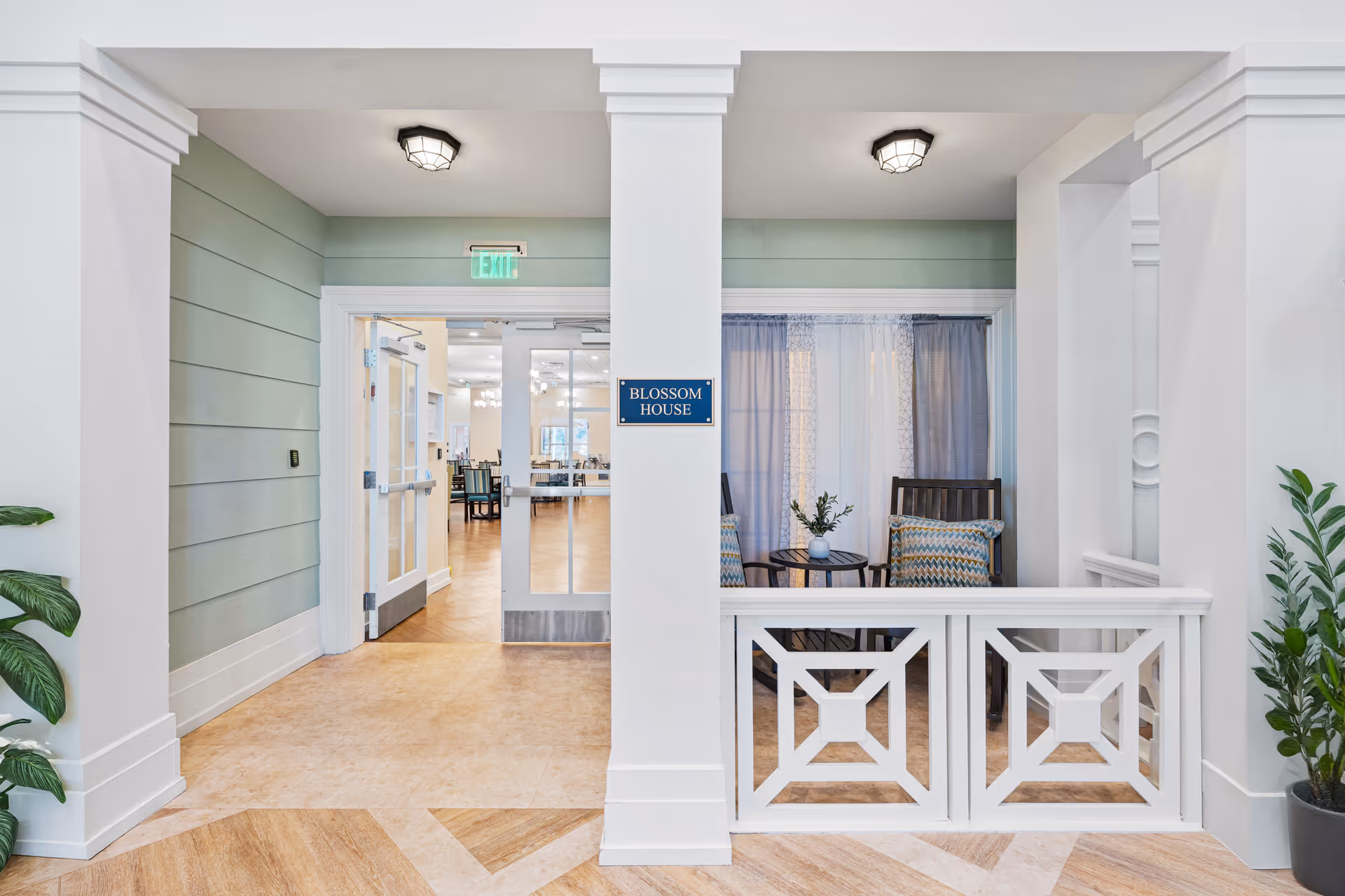Interior view of a senior living facility hallway with light green walls and white trim. There is a sign on a central column that reads 'Blossom House.' To the right, there is a small seating area with two wooden chairs, patterned cushions, a small round table with a plant, and sheer curtains. Through double glass doors ahead, a dining area with tables and chairs is visible. Potted plants are placed on either side of the hallway.